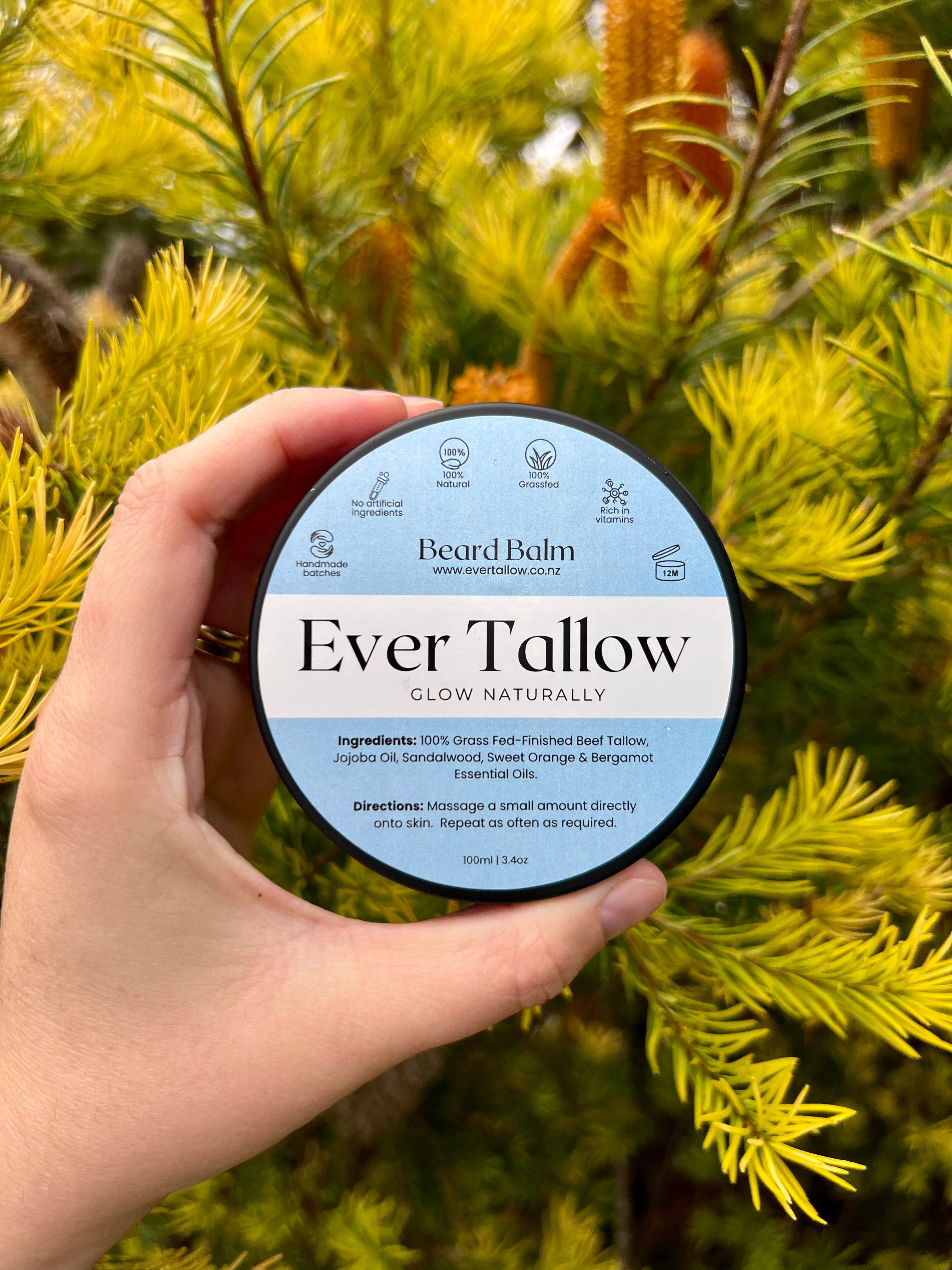 Hand holding a container of 'Ever Tallow' Beard Balm against a green foliage background