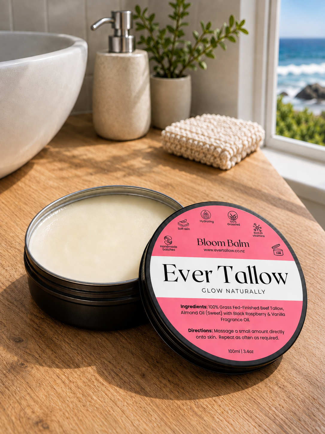 An open tin of Ever Tallow Glow on a wooden bathroom counter beside a sink, soap dispenser, plant, and a window view of the ocean. 
