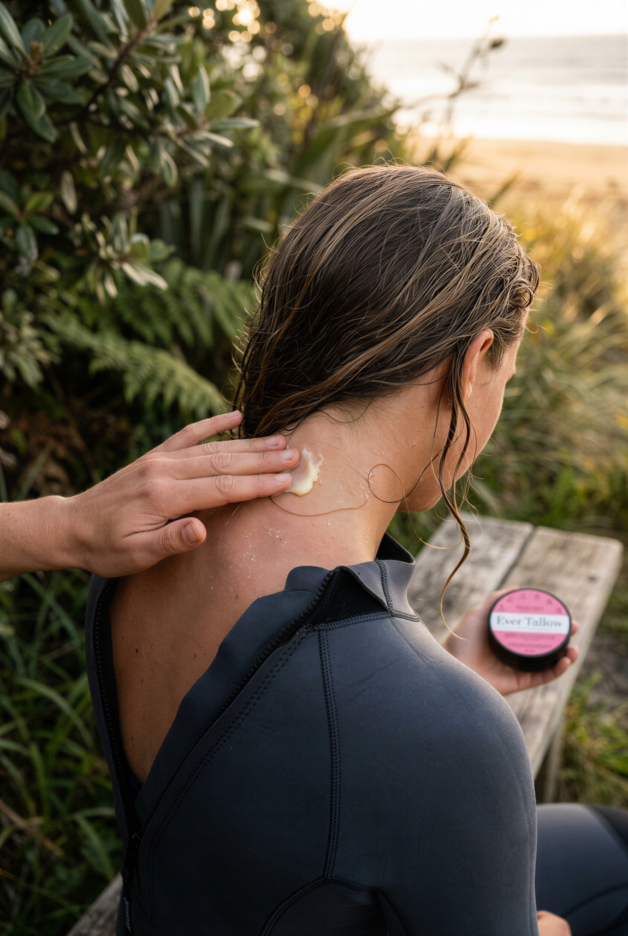 A woman in a black wetsuit sits outdoors by the beach, receiving tallow balm applied to her neck from another person's hand. She holds bloom balm in her hand.