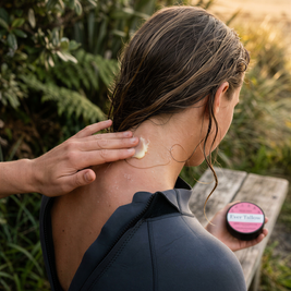 A woman in a black wetsuit sits outdoors by the beach, receiving tallow balm applied to her neck from another person's hand. She holds bloom balm in her hand.
