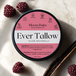A round metal tin of Bloom Balm Ever Tallow fragrance oil sits on a light marble surface, surrounded by whole raspberries and a vanilla bean. The lid is pink and white with black text, and the tin has a black rim.