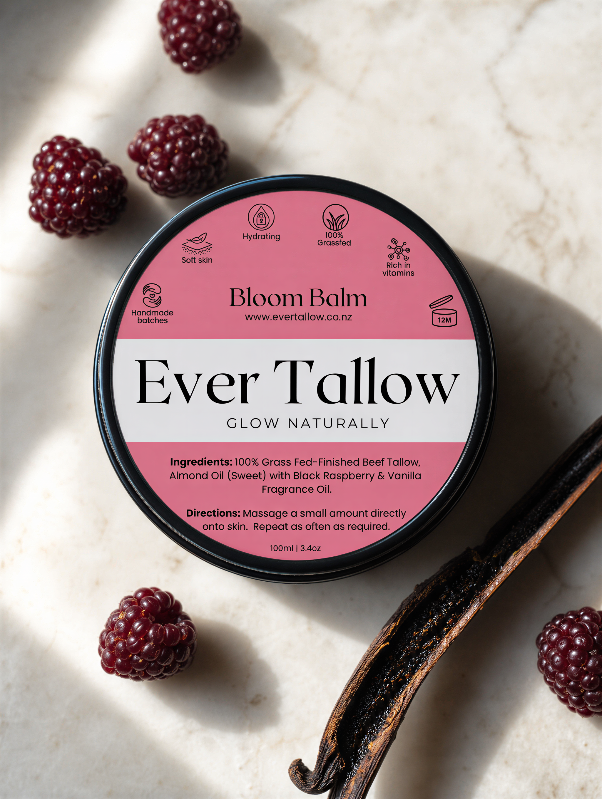 A round metal tin of Bloom Balm Ever Tallow fragrance oil sits on a light marble surface, surrounded by whole raspberries and a vanilla bean. The lid is pink and white with black text, and the tin has a black rim.