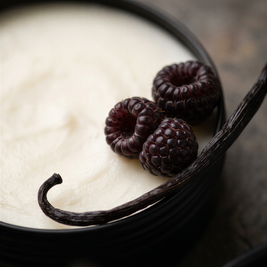 A close-up of a creamy off-white tallow balm in a tin, with three dark raspberries and a vanilla bean on a dark surface.