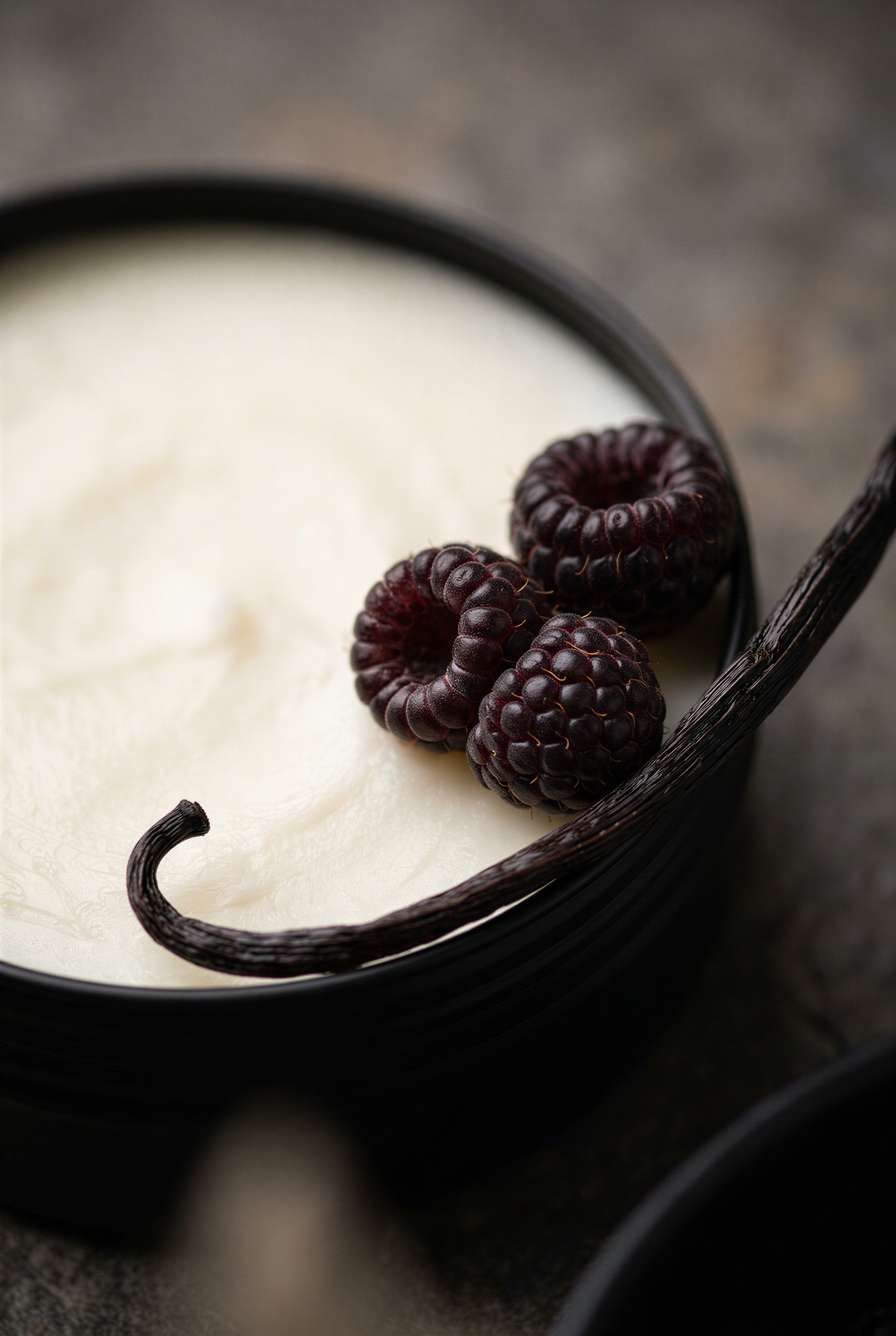 A close-up of a creamy off-white tallow balm in a tin, with three dark raspberries and a vanilla bean on a dark surface.
