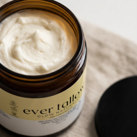 An open amber glass jar filled with whipped tallow with its black lid resting beside it on a beige textured fabric surface.