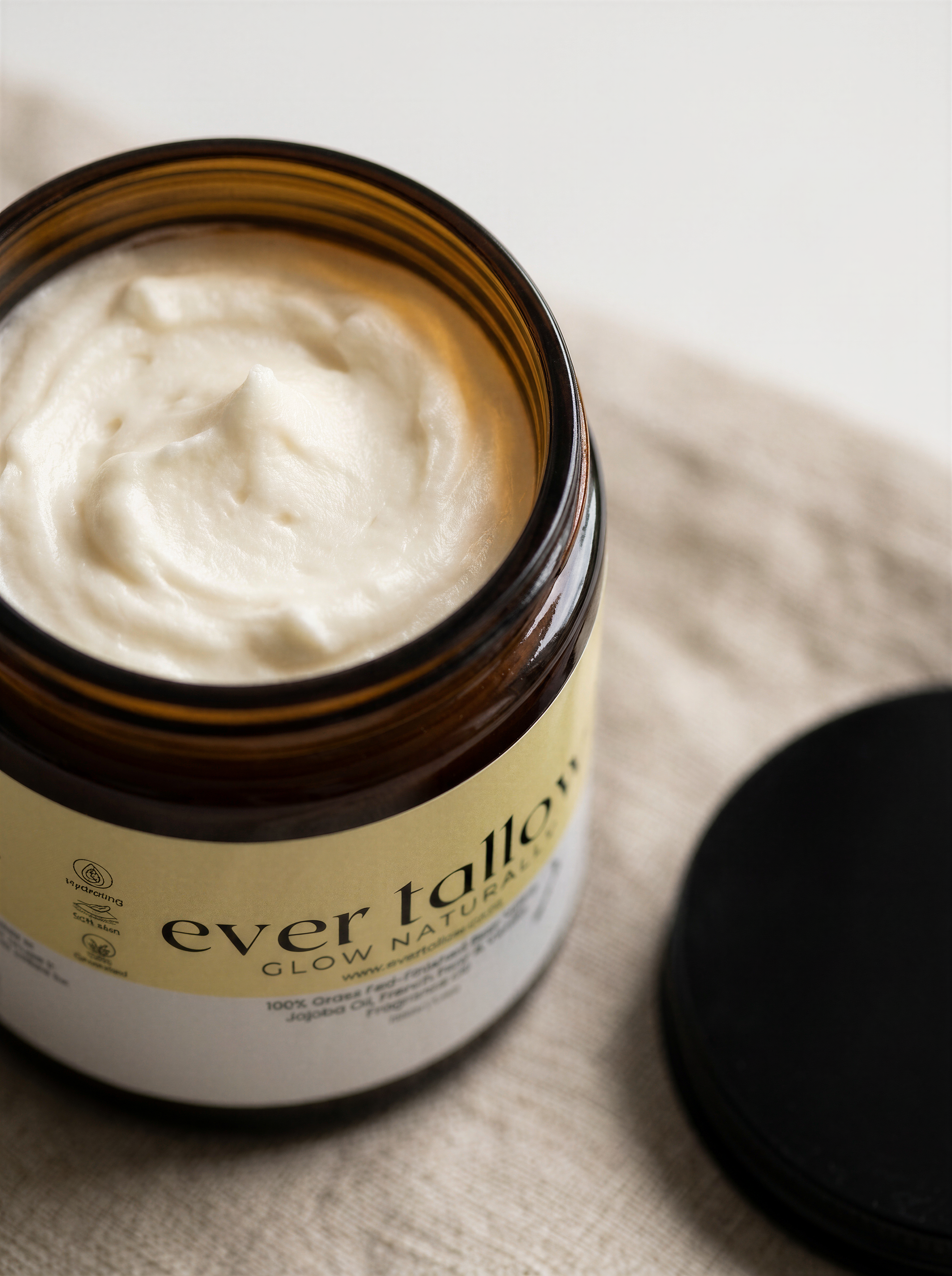 An open amber glass jar filled with whipped tallow with its black lid resting beside it on a beige textured fabric surface.