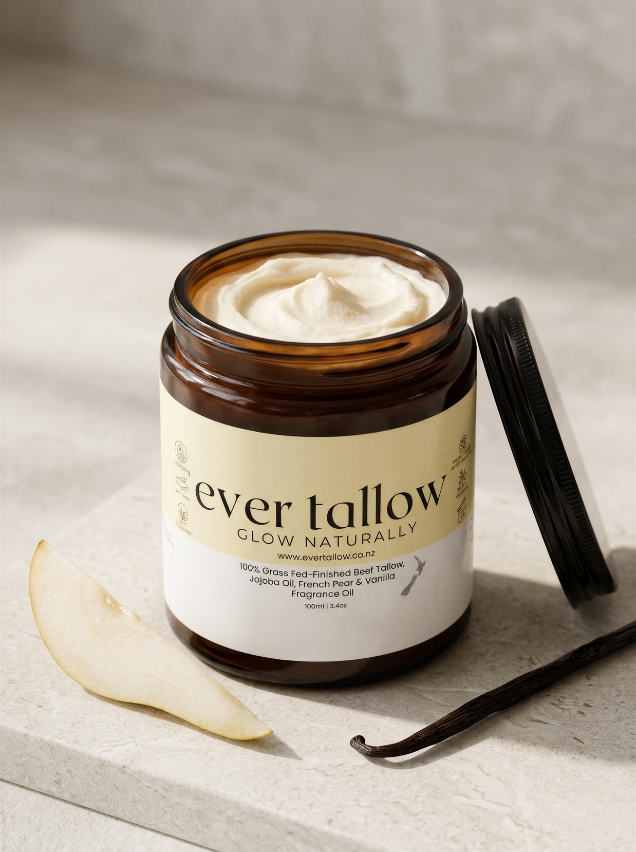 An open amber glass jar of Ever Tallow Glow Naturally body butter with French Pear and Vanilla whipped cream visible inside, set on a light stone surface with a vanilla bean and a pear slice nearby. The lid rests beside the jar.