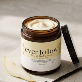 An open amber glass jar of Ever Tallow Glow Naturally body butter with French Pear and Vanilla whipped cream visible inside, set on a light stone surface with a vanilla bean and a pear slice nearby. The lid rests beside the jar.