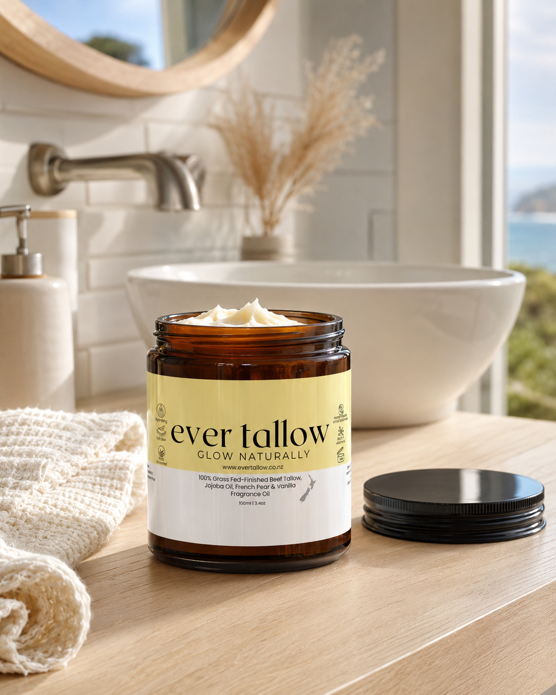 An amber glass jar of French Pear and Vanilla body butter sits on a light wood countertop in a bright bathroom setting, with its black lid placed beside it. A white sink, towel, and window view are visible in the background.