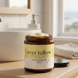 A jar of Ever Tallow Glow Naturally French Pear and Vanilla butter sits on a light wood vanity counter in a bright bathroom setting, with its lid placed beside it.