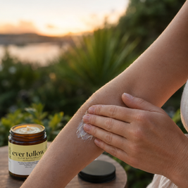A person applying a French Pear and Vanilla Whipped Tallow cream from an amber glass jar outdoors at sunset, with a blurred natural background and the jar resting on a wooden table.