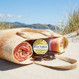 Fruity Balm in an open beach bag on pale sand. Soft woven beach bag texture, natural fibres, light summer colours.