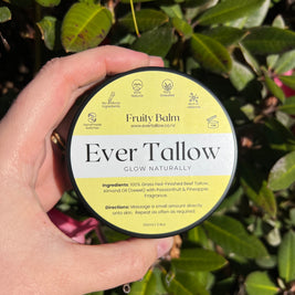 Hand holding a container of 'Fruity Tallow' balm against a green leafy background