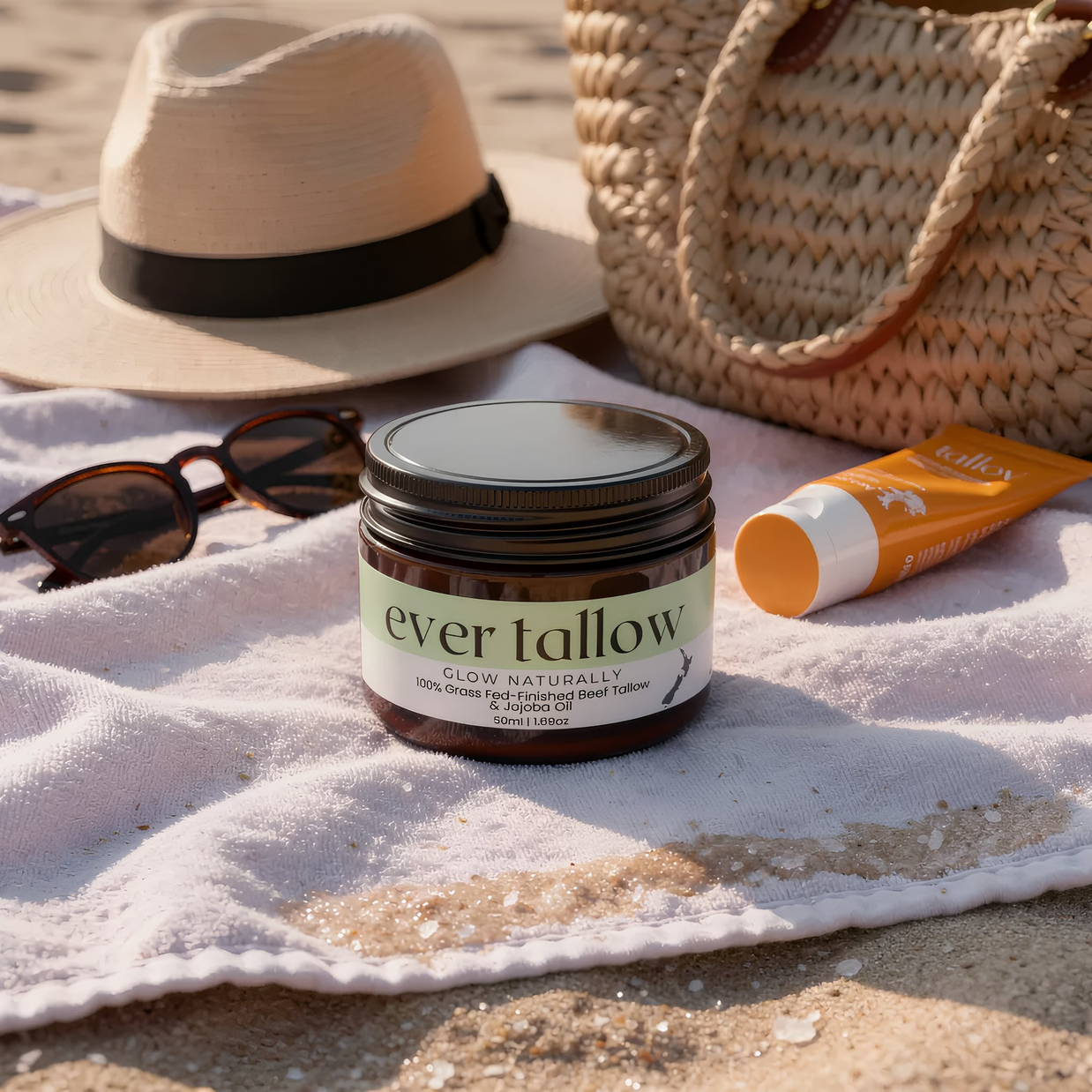 Jar of 'ever tallow' product on a beach towel with a hat, sunglasses, and sunscreen.