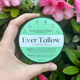 Hand holding a container of Ever Tallow Kawakawa Balm with pink flowers in the background