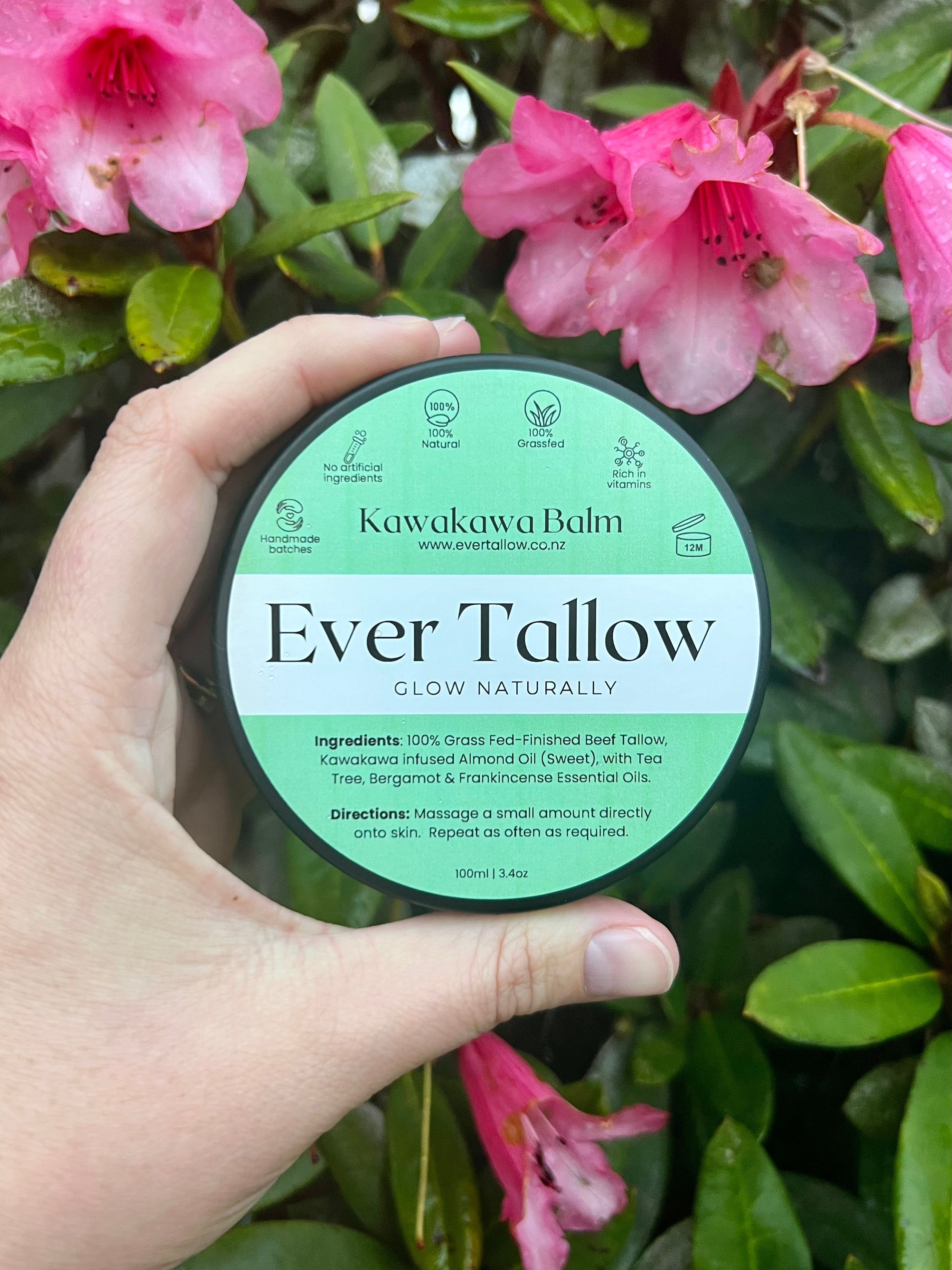 Hand holding a container of Ever Tallow Kawakawa Balm with pink flowers in the background
