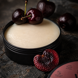 A black round tin filled with a pale creamy velvet balm owith whole and halved dark red cherries on a dark textured surface. Part of the Ever Tallow label is visible in the lower right corner.