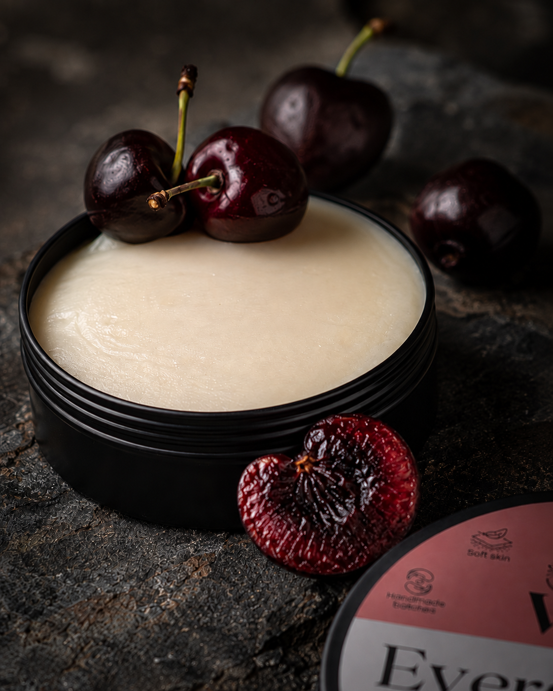 A black round tin filled with a pale creamy velvet balm owith whole and halved dark red cherries on a dark textured surface. Part of the Ever Tallow label is visible in the lower right corner.