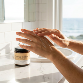 A person applying cream to hands with a jar of vanilla-infused tallow cream on a marble surface.