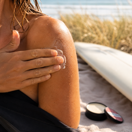 A close-up lifestyle shot of a woman at the beach applying Velvet Balm to her shoulder. A small black tin and its lid sit on a towel nearby, with a surfboard and ocean dunes in the background.