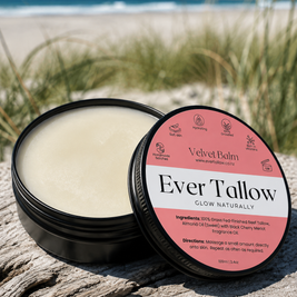 An open round black tin of Ever Tallow Velvet balm sits on weathered wood with a beach and dune grasses blurred in the background. The lid is placed beside the tin.
