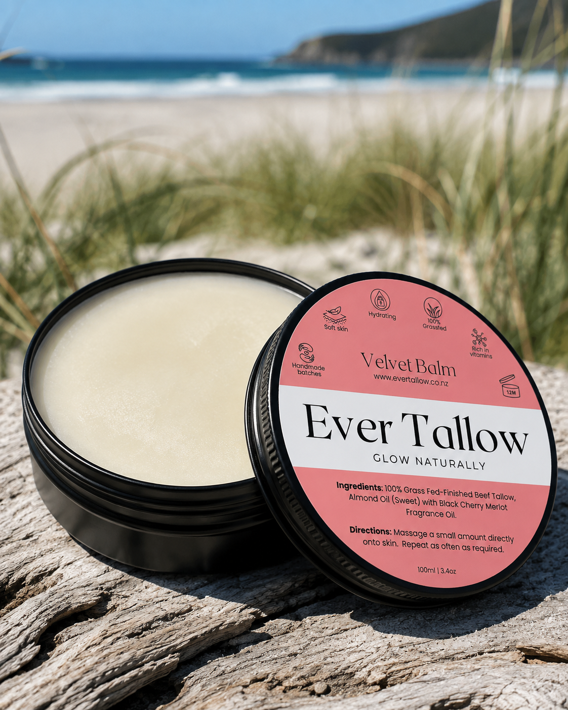 An open round black tin of Ever Tallow Velvet balm sits on weathered wood with a beach and dune grasses blurred in the background. The lid is placed beside the tin.
