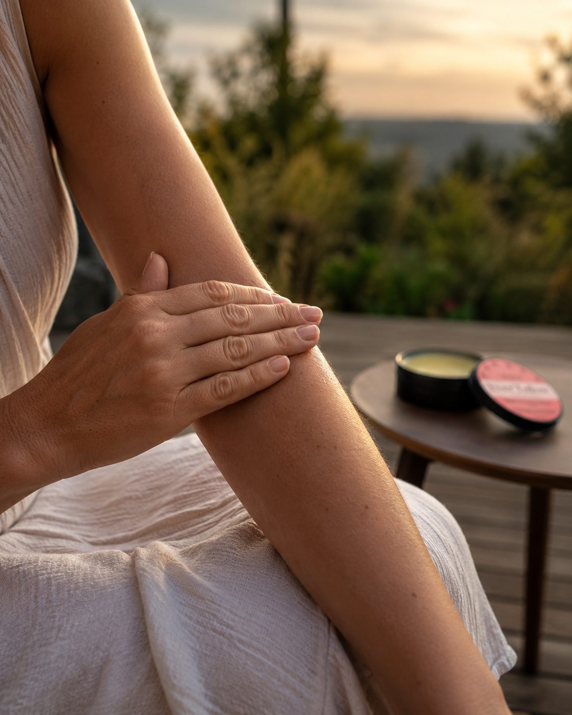 A close-up lifestyle shot of a person applying Velvet Balm from an open round tin onto their forearm, set against an outdoor scenic background at sunset.
