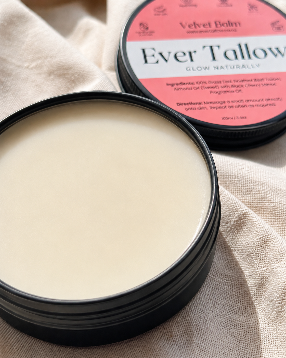 An open round black tin of Ever Tallow Velvet Balm showing the creamy off-white solid product inside, with the branded lid resting behind it on a beige fabric background.