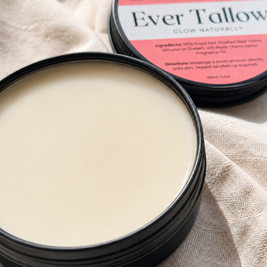 An open round black tin of Ever Tallow Velvet Balm showing the creamy off-white solid product inside, with the branded lid resting behind it on a beige fabric background.